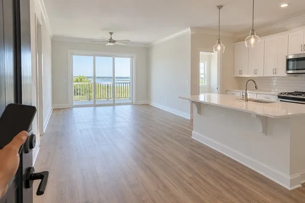 Empty apartment with an open kitchen, wood-look floors, and balcony doors during an in-person tour.